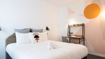 Een netjes opgemaakt bed met witte lakens en een opgerolde handdoek, een nachtkastje met een lamp, en een wand met geometrische patronen en een grote ronde lamp.