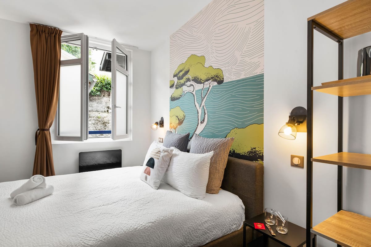 Hotellihuone, jossa on sänky, ikkuna verhoilla, seinämaalaus, yöpöytä laseineen ja hylly.
