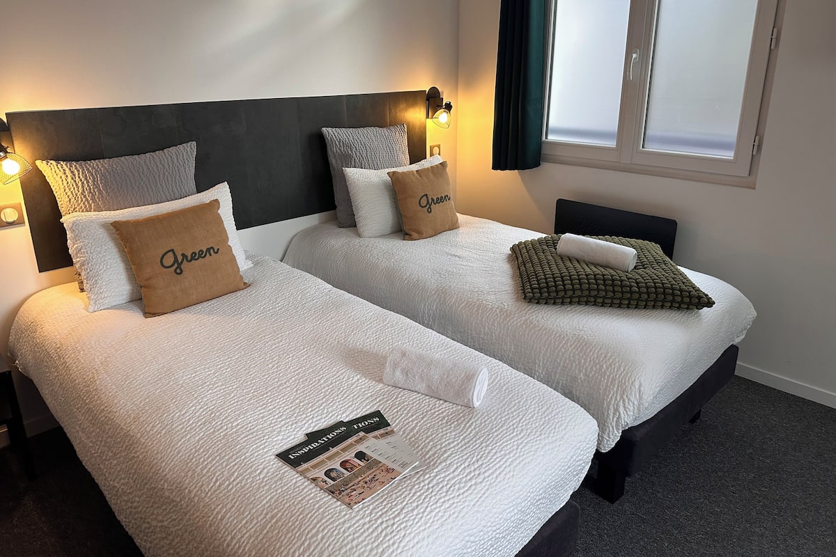 Hotellihuoneessa on kaksi sänkyä, joissa on valkoiset lakanat ja useita tyynyjä, mukaan lukien vihreä tyyny. Huoneessa on ikkuna, jossa on verhot, seinään kiinnitetty valaisin ja sängyllä lehti.