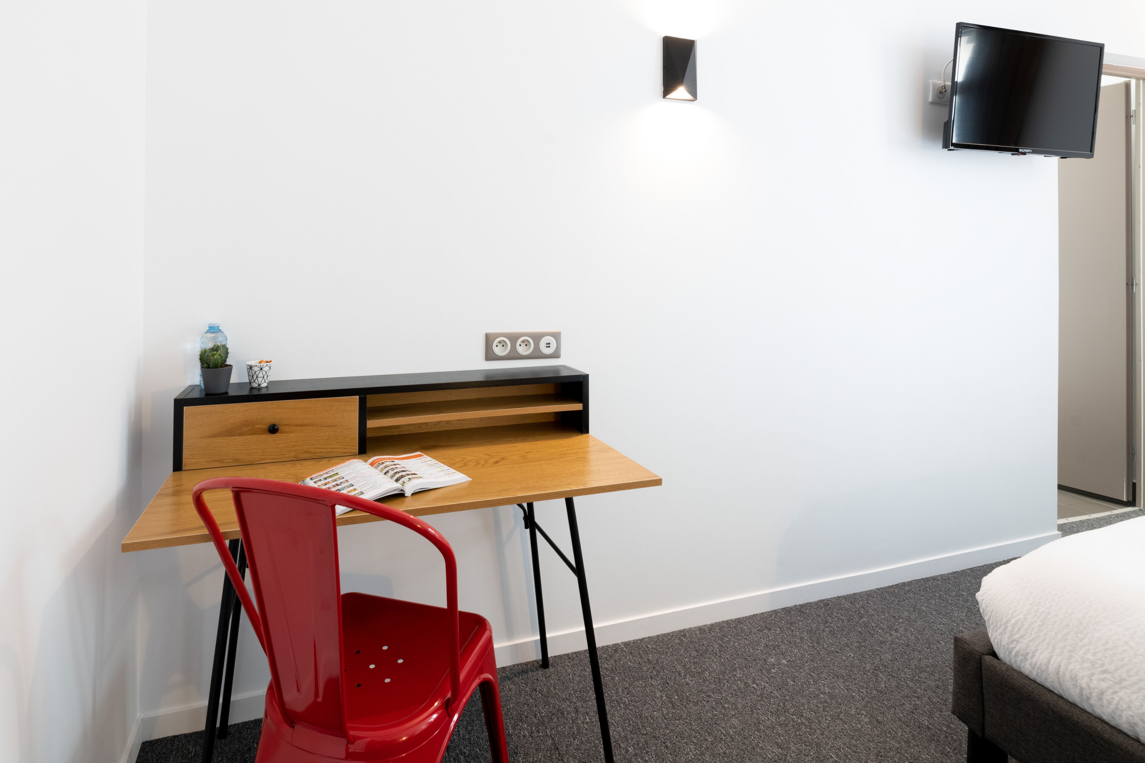 Habitación de hotel moderna con un escritorio de madera, una silla roja, un televisor de pantalla plana montado en la pared y una cama con edredón blanco.