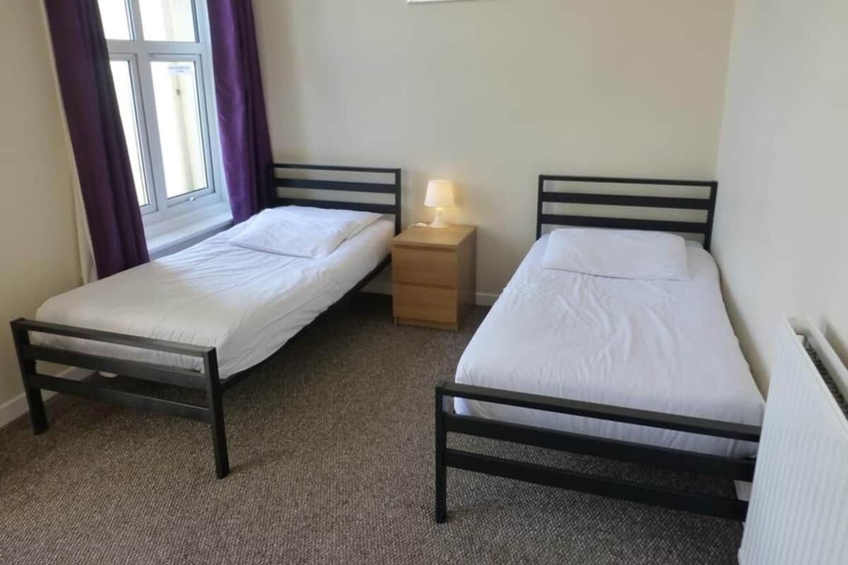 A room with two single beds, a nightstand with a lamp, a window with purple curtains, and a radiator.