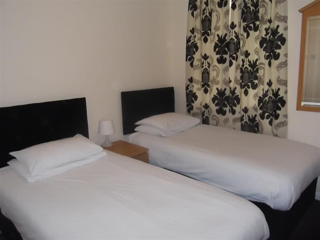 Ein Hotelzimmer mit zwei Einzelbetten, einem Nachttisch, einer Lampe und gemusterten Vorhängen.