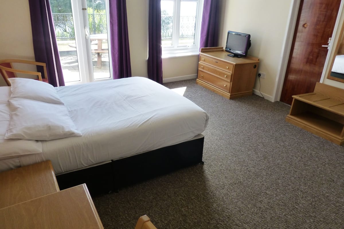 A hotel room with a bed, a wooden dresser, a television on a stand, and a window with curtains.