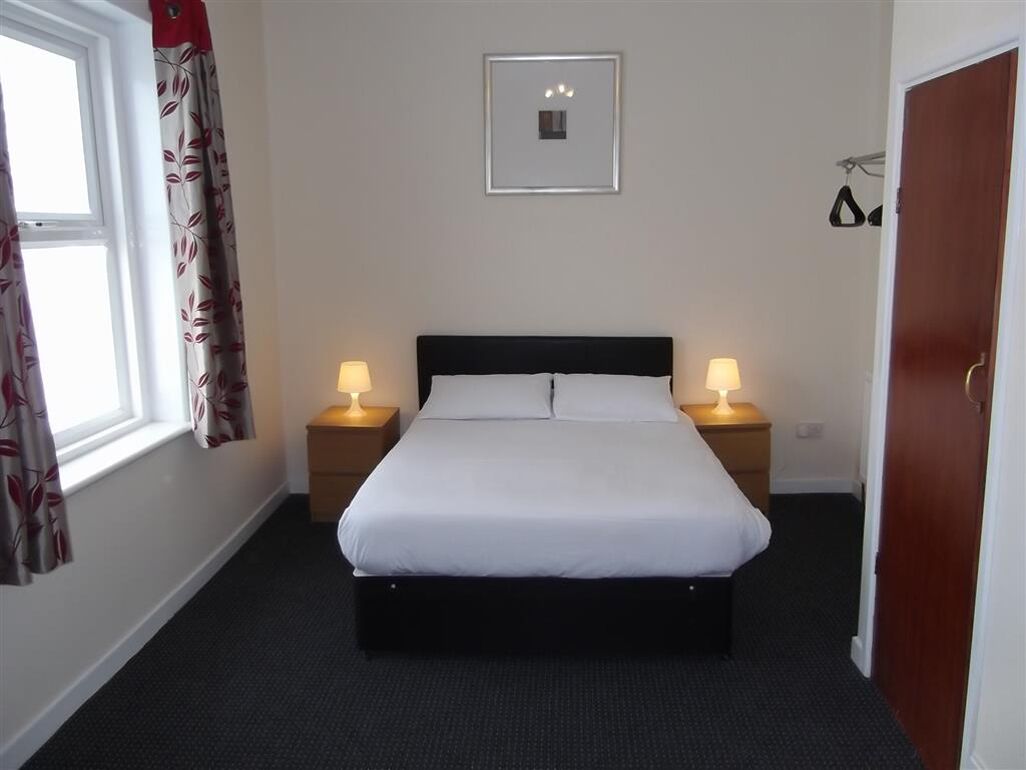 A neatly made bed in a hotel room with a window, two bedside lamps, and a framed picture on the wall.