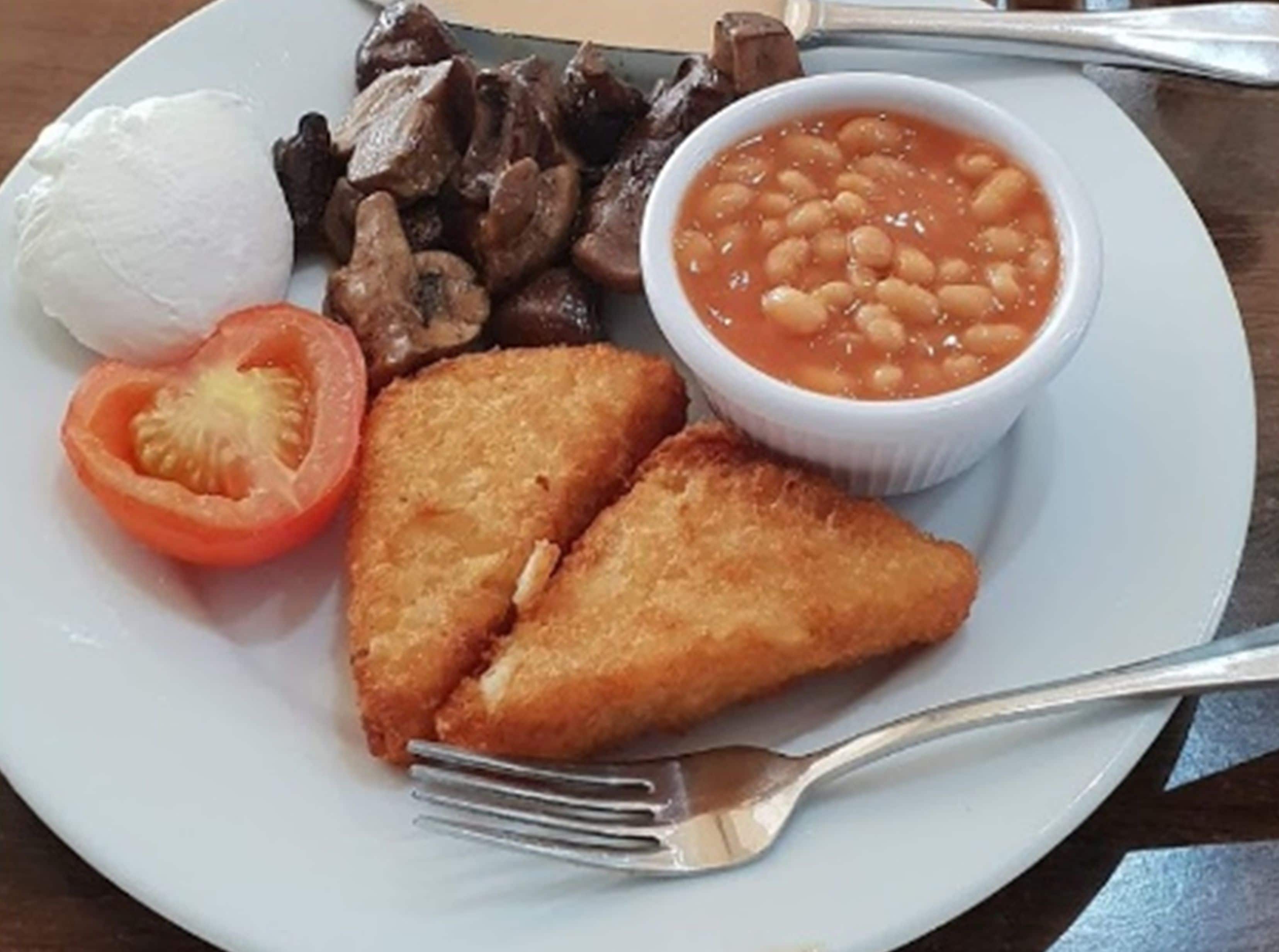 Un plato con un huevo cocido, champiñones fritos, frijoles horneados, una rodaja de tomate y dos preparaciones fritas.