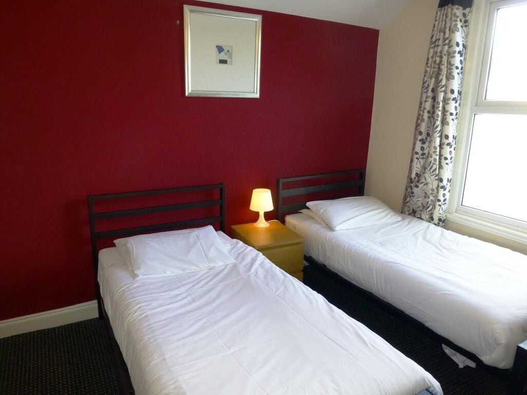 A hotel room with two single beds, a nightstand with a lamp, a framed picture on the wall, and a window with curtains.