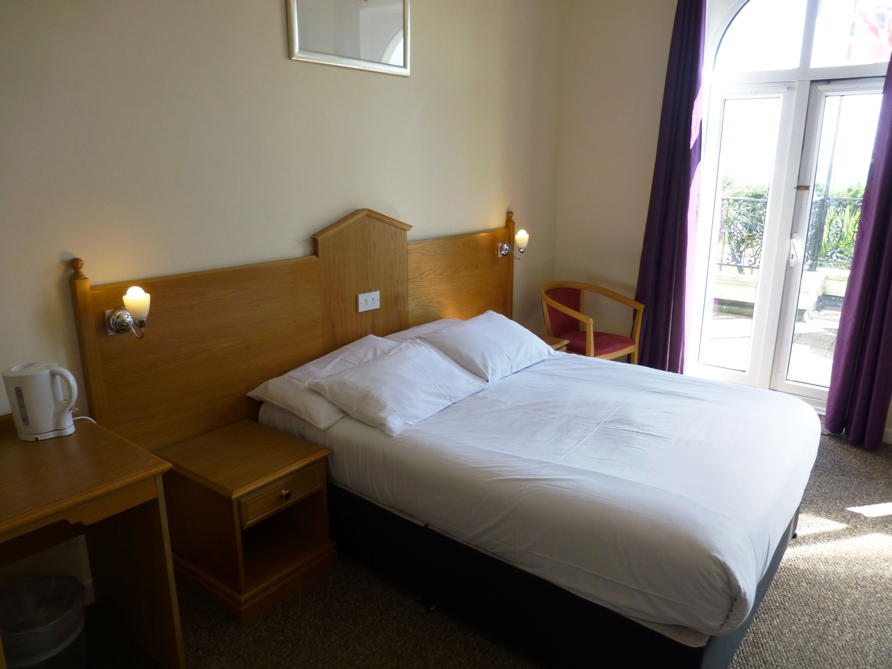 A hotel room with a wooden bed, a nightstand with a kettle, a chair, and a view of the outdoors through a sliding door.