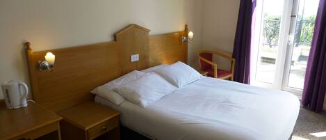 A hotel room with a wooden bed, a nightstand with a kettle, a chair, and a view of the outdoors through a sliding door.