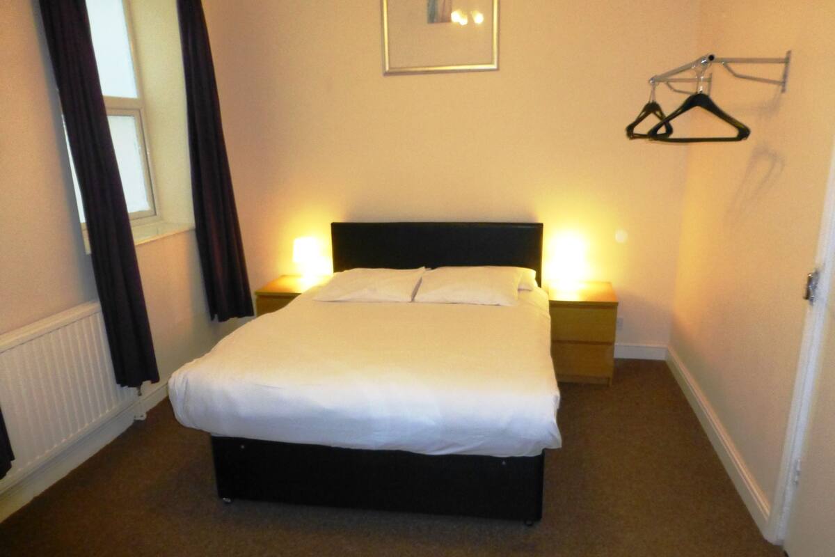 A neatly made bed in a hotel room with a headboard, two bedside tables with lamps, a window with curtains, and a coat rack.