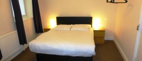 A neatly made bed in a hotel room with a headboard, two bedside tables with lamps, a window with curtains, and a coat rack.