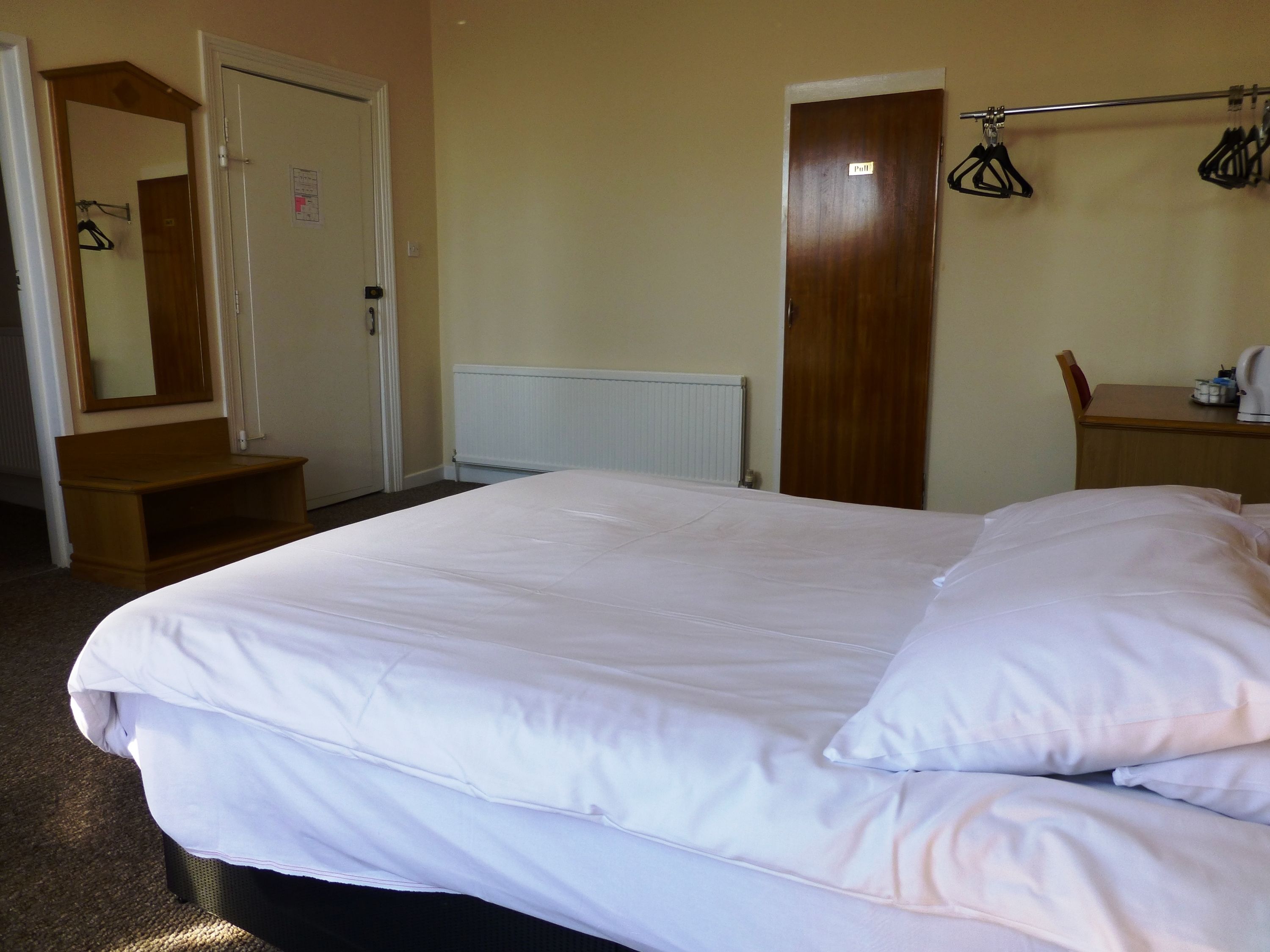 A hotel room with a large bed, a wooden bench, a radiator, a wardrobe, a mirror, and a coat rack.