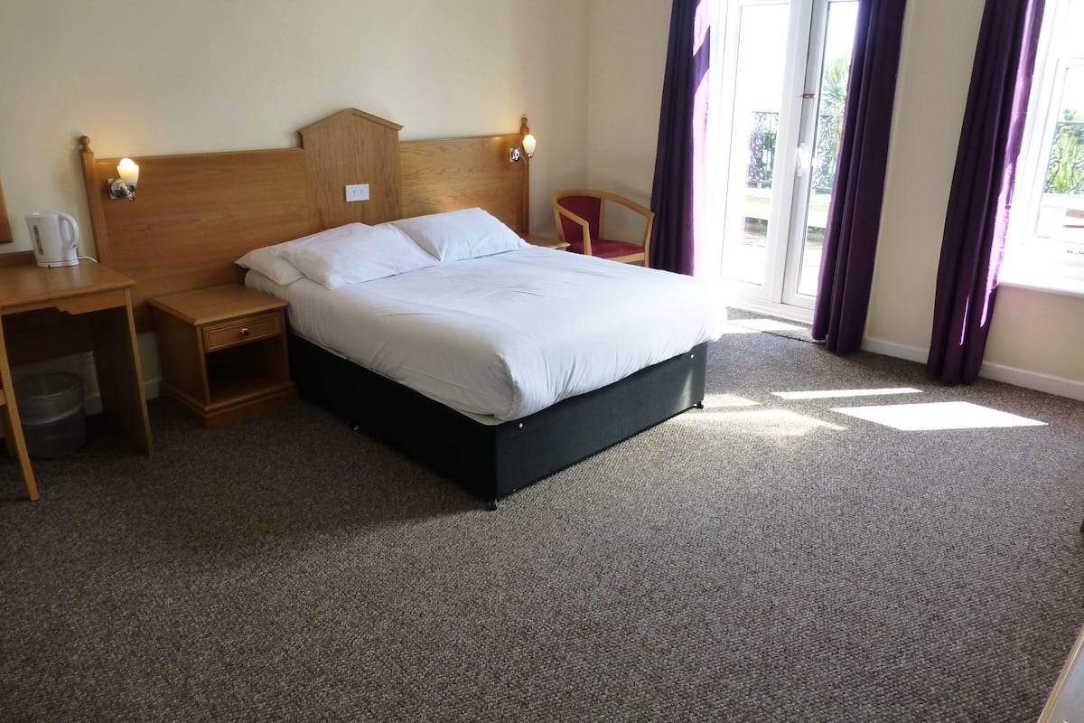 A hotel room with a bed, bedside table, lamp, and a view of the outdoors.