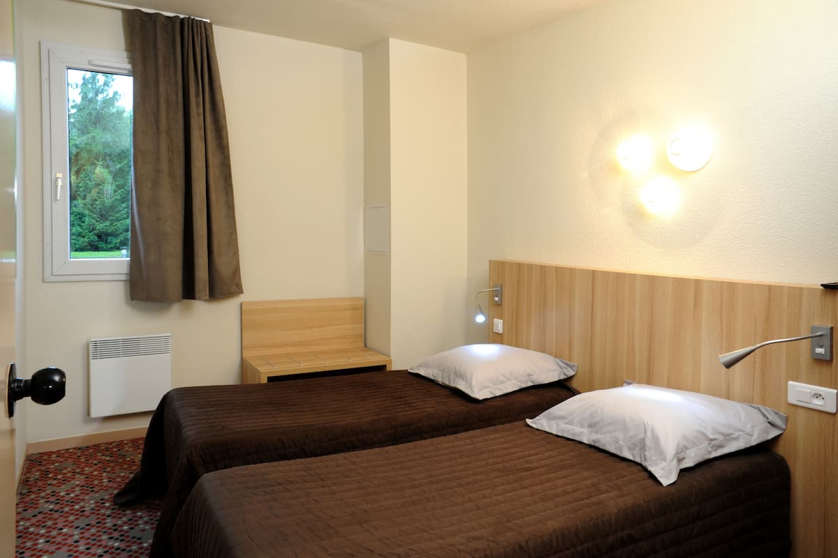 A hotel room with two beds, a wooden headboard, a window with curtains, a wall-mounted lamp, and a radiator.
