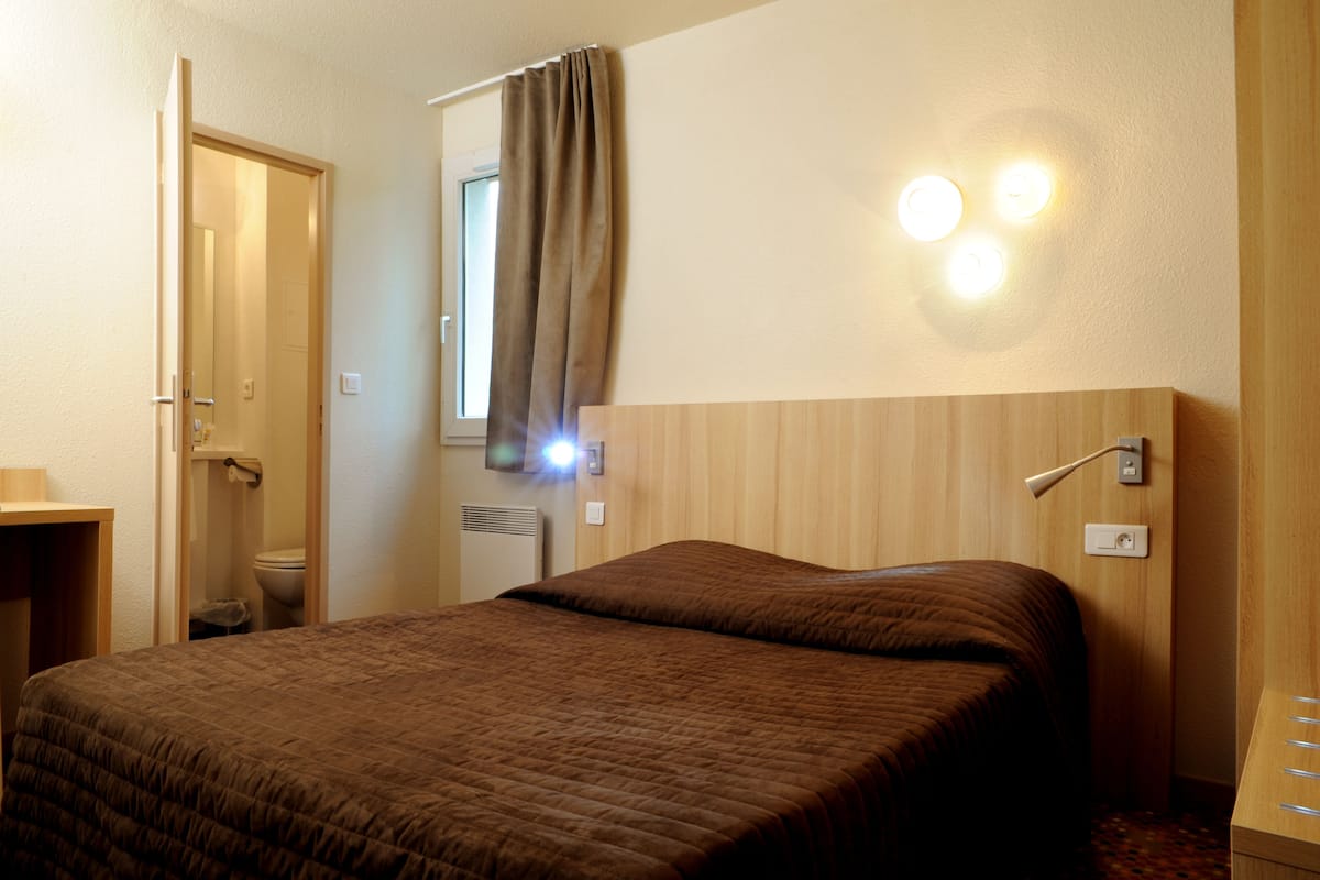 A hotel room with a bed, a bedside table, a window with curtains, and wall-mounted lights.