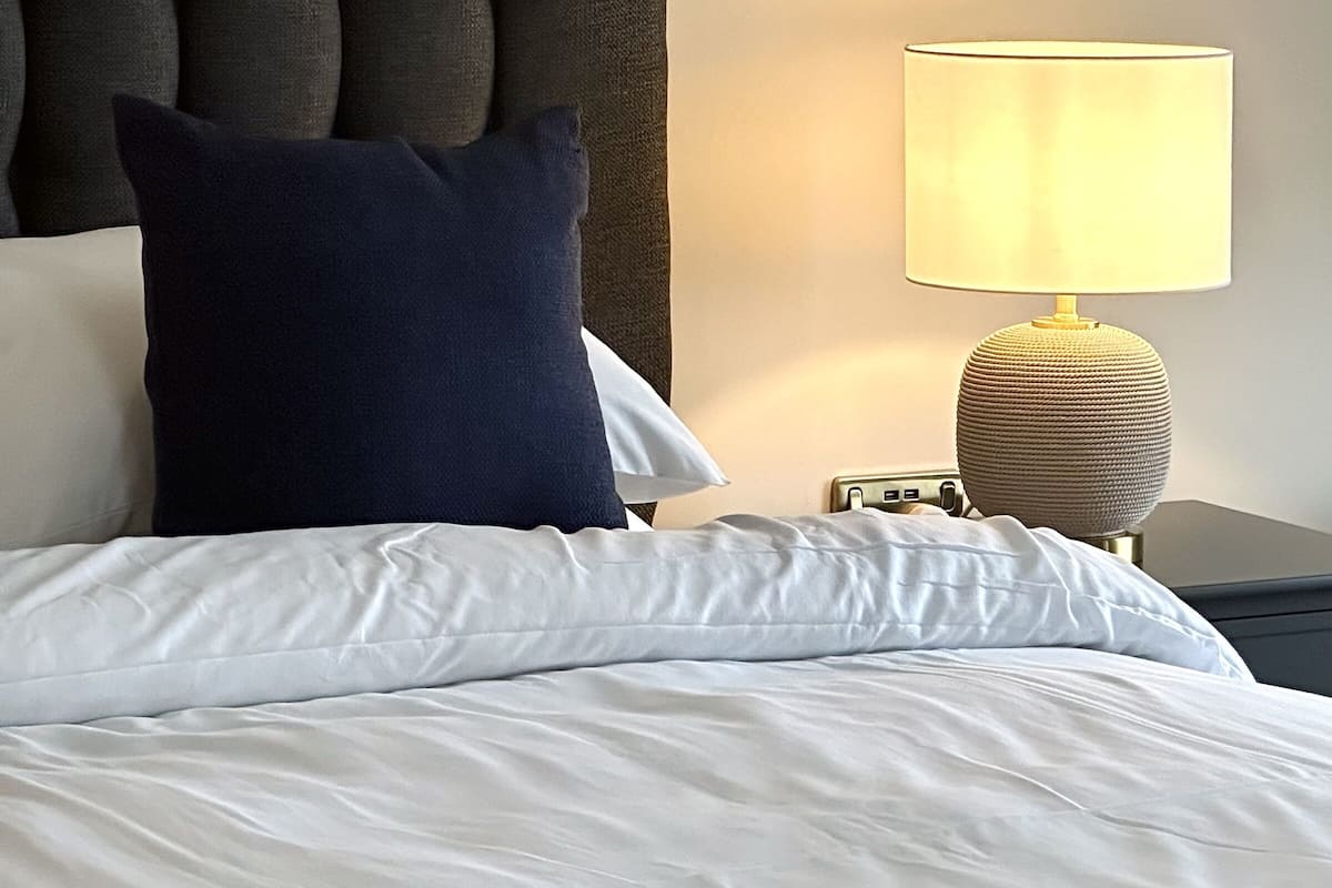 A neatly made bed with white linens and a dark blue pillow, a round lamp with a textured base, and a tufted headboard.