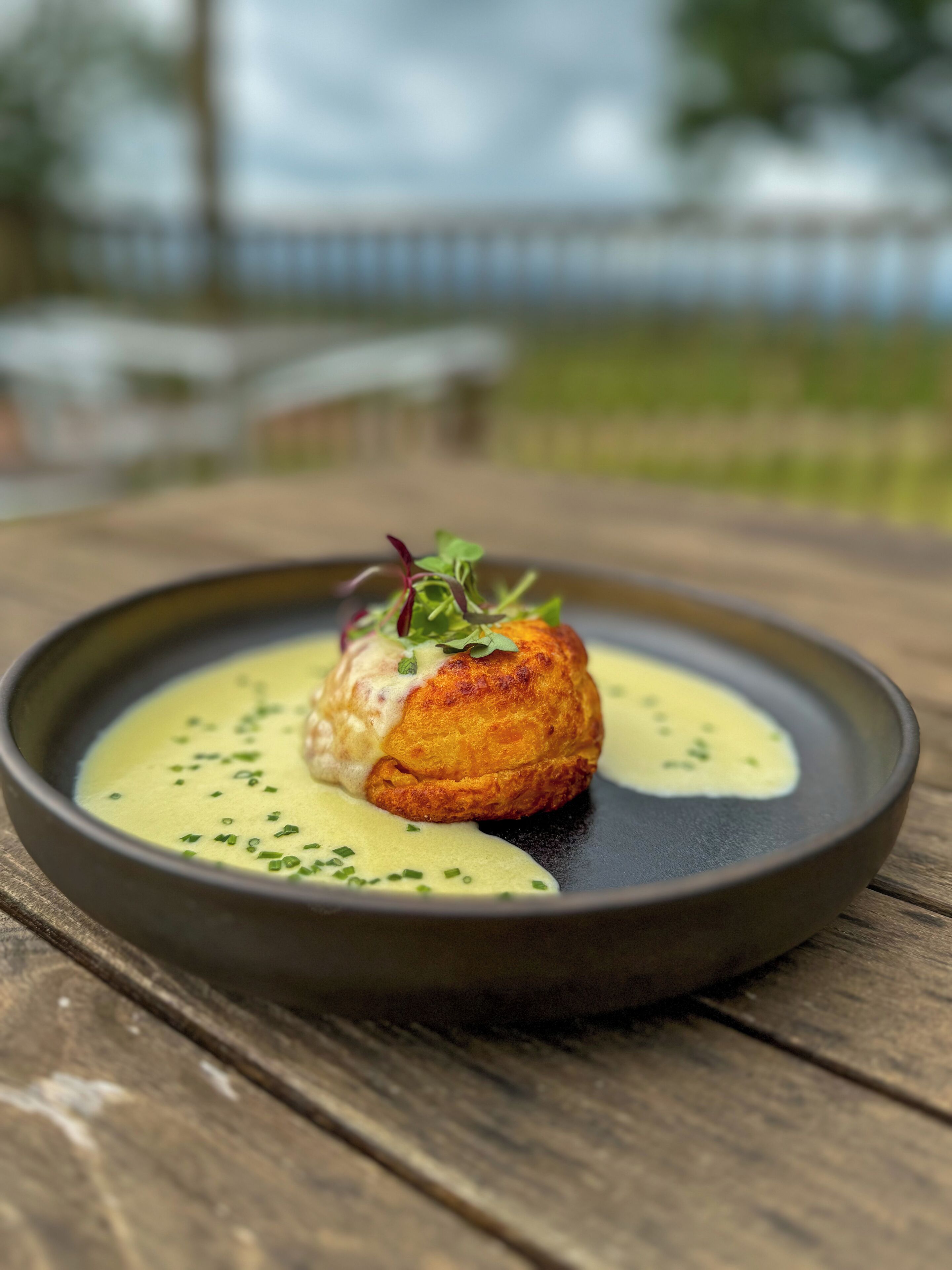 A dish with a golden-brown pastry on a black plate, garnished with green herbs, served on a wooden table.