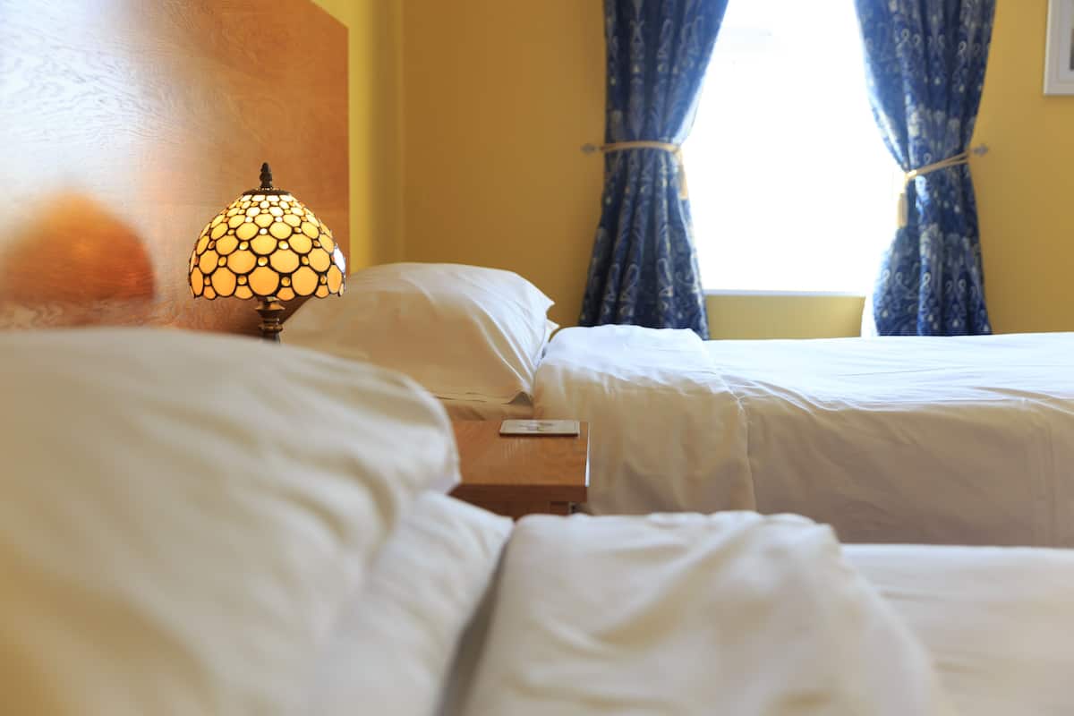 A hotel room with two beds, a bedside table with a lamp, and a window with curtains.