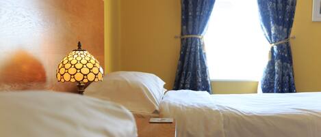 Een hotelkamer met twee bedden, een nachtkastje met een lamp en een raam met gordijnen.