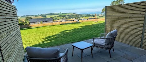 An outdoor patio with two wicker chairs and a small table, overlooking a green field and distant hills.