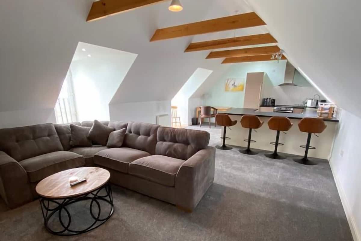 A loft-style living area with a large L-shaped sectional sofa, a round coffee table, and a kitchen in the background.