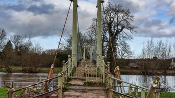 Un puente con estructura metálica y escalones de madera que conducen a un portón, rodeado de césped y una masa de agua.