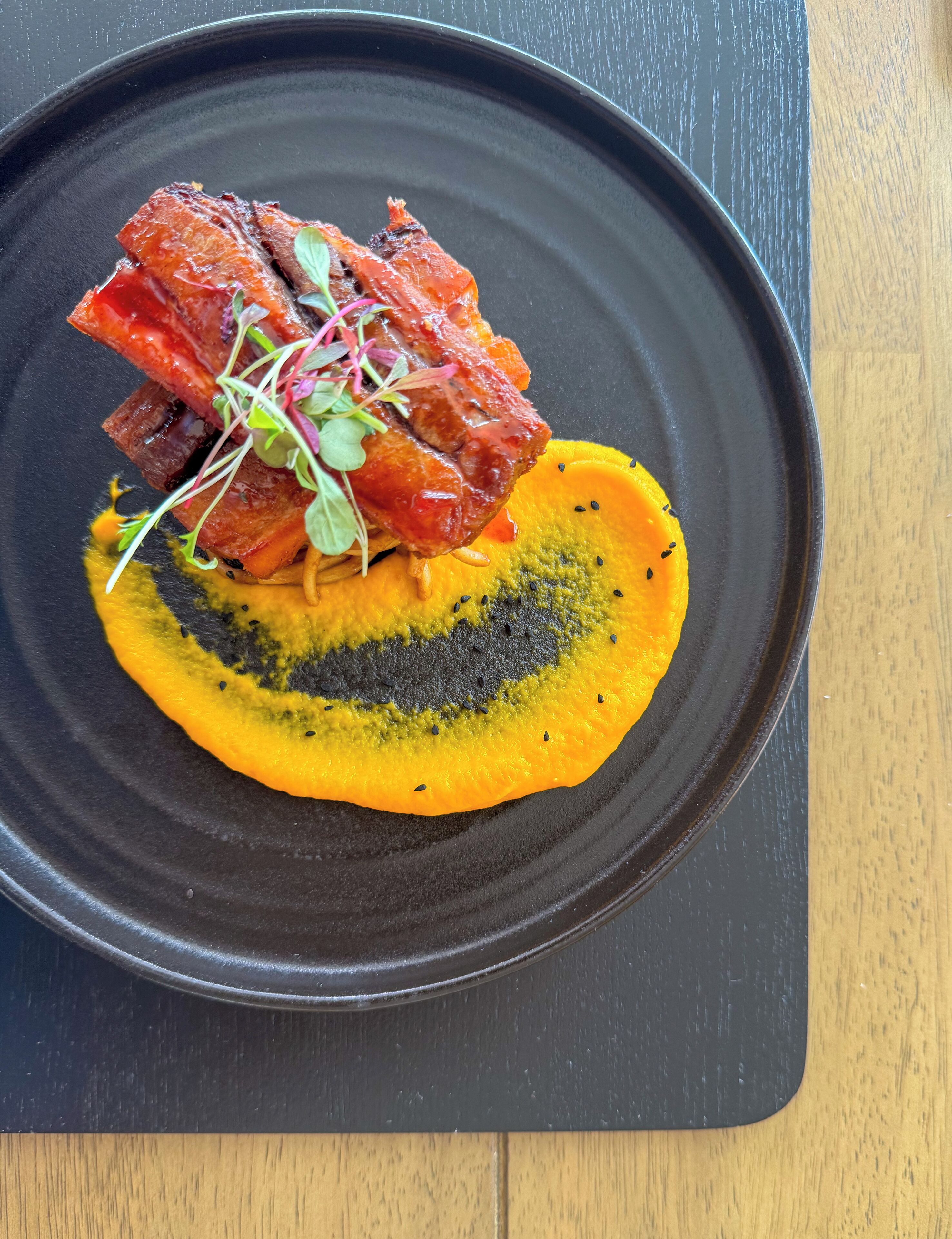 Un piatto nero con un piatto di cibo, tra cui sembra esserci carne e una fetta di frutto giallo, forse un mango, su un tavolo di legno.