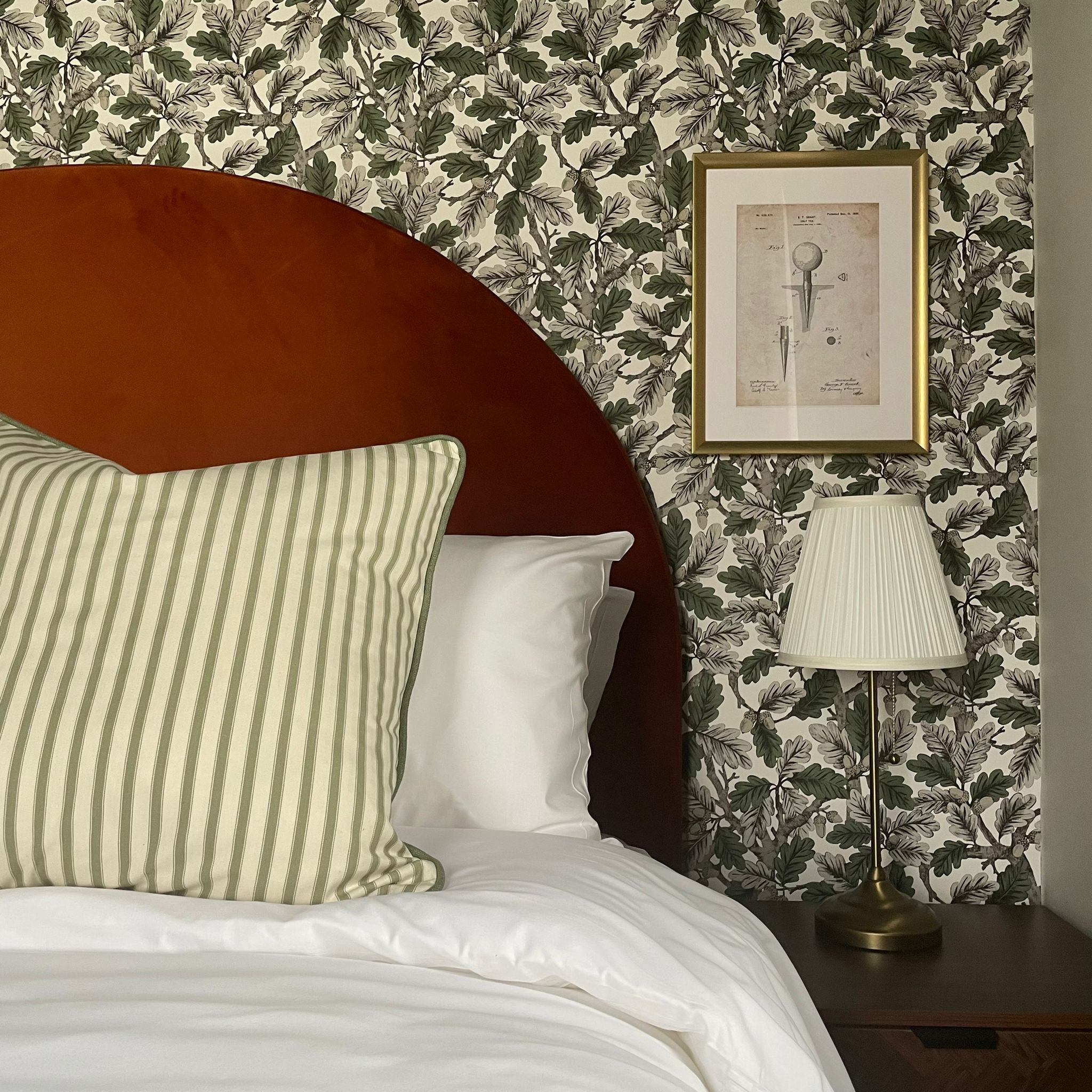 Cama bem arrumada com travesseiro listrado e um travesseiro branco, cabeceira redonda de madeira, impressão botânica emoldurada na parede e abajur em uma mesa de cabeceira.