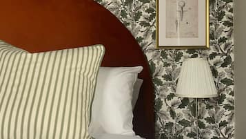A neatly made bed with a striped pillow and a white pillow, a round wooden headboard, a framed botanical print on the wall, and a lamp on a bedside table.