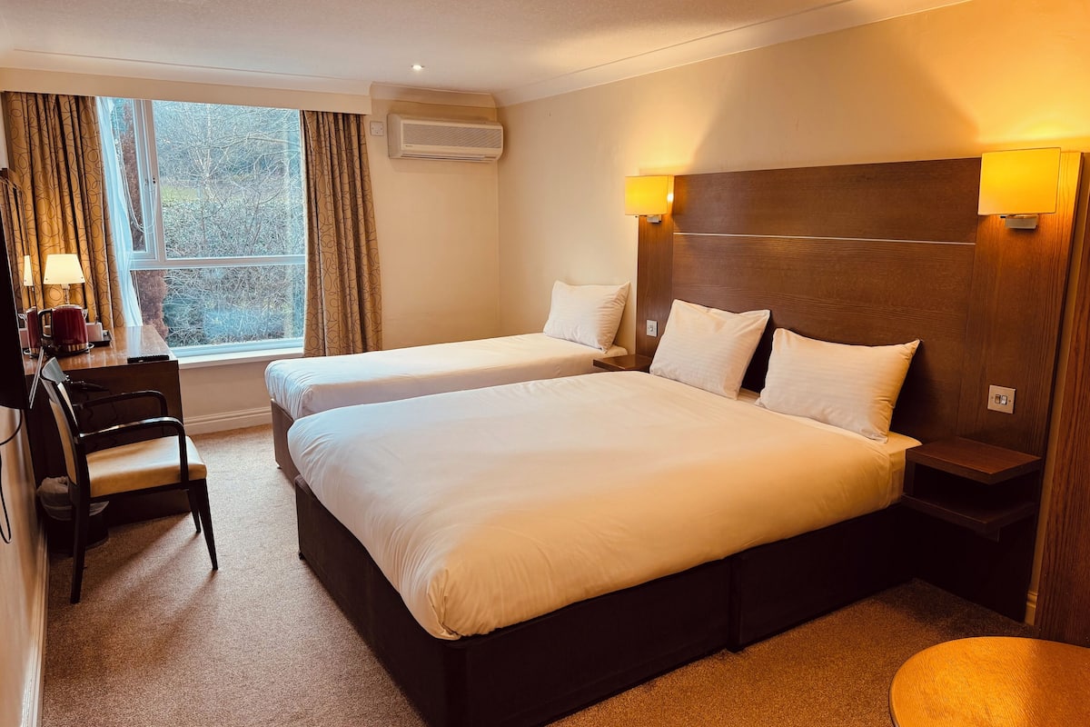 A hotel room with two beds, a desk, a chair, a lamp, and a window with curtains.