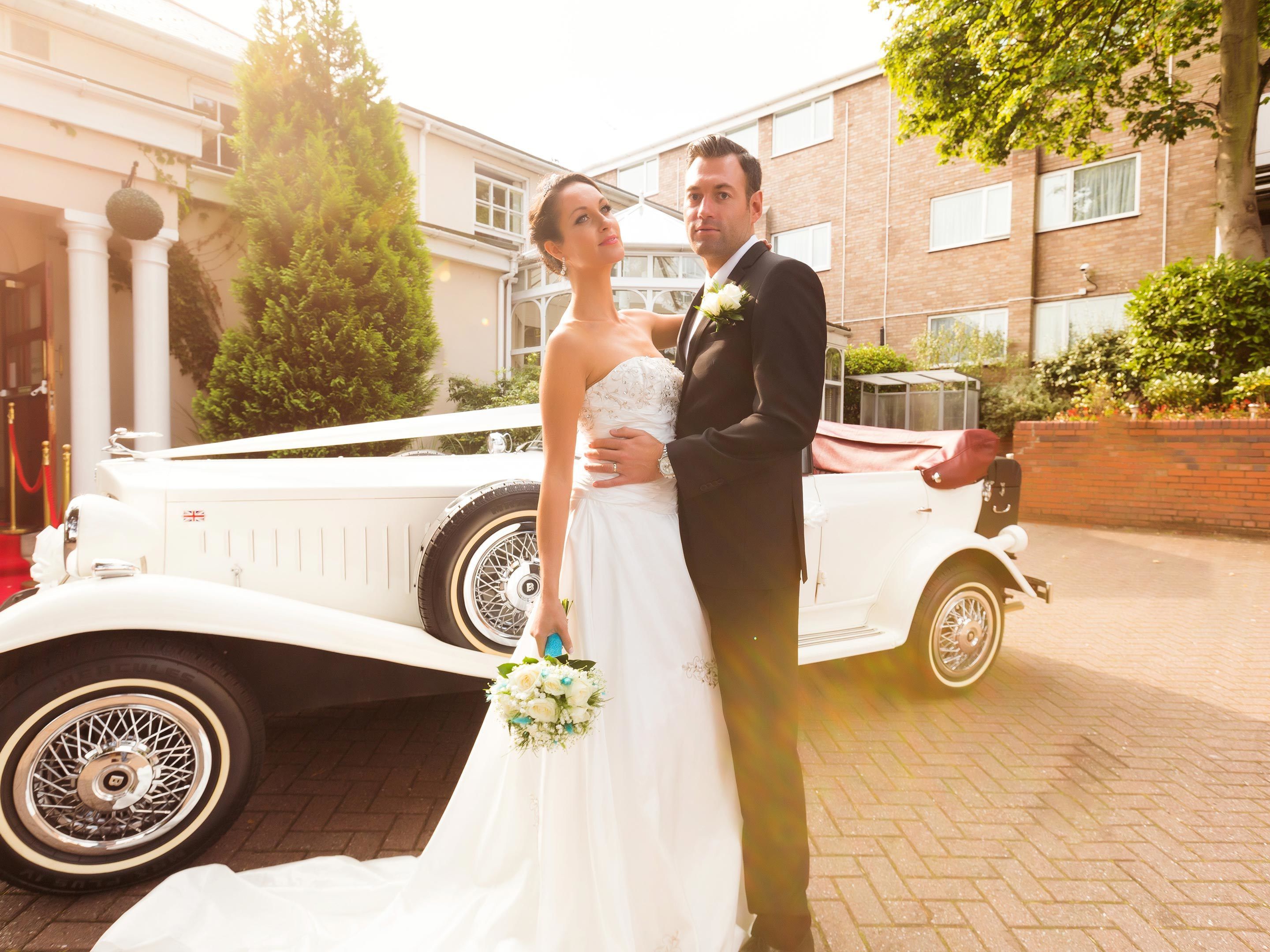A wedding couple standing in front of a vintage car with a building in the background.