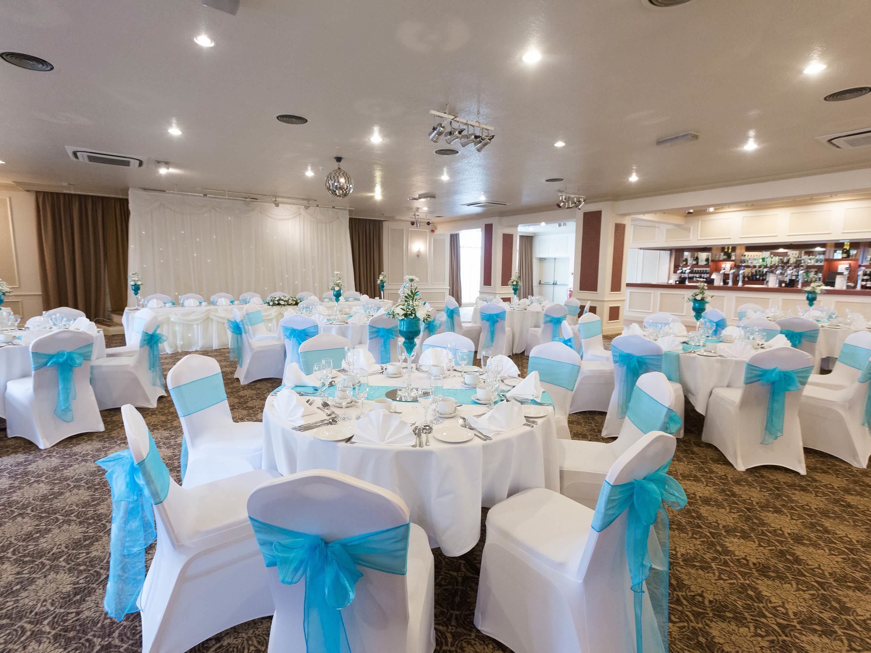 A banquet hall with round tables set for a formal event, white chairs with blue sashes, and a bar area in the background.