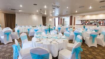A banquet hall with round tables set for a formal event, white chairs with blue sashes, and a bar area in the background.