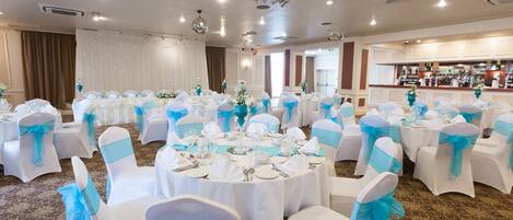 A banquet hall with round tables set for a formal event, white chairs with blue sashes, and a bar area in the background.