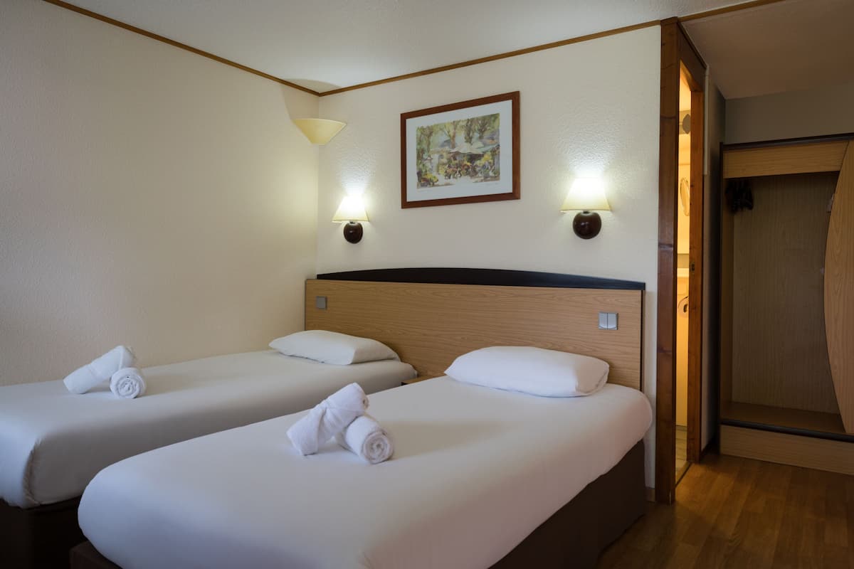 A hotel room with two single beds, each with a rolled towel, a wooden headboard, and two wall-mounted lamps.