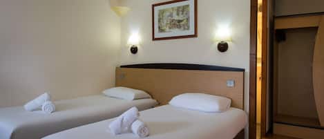 A hotel room with two single beds, each with a rolled towel, a wooden headboard, and two wall-mounted lamps.