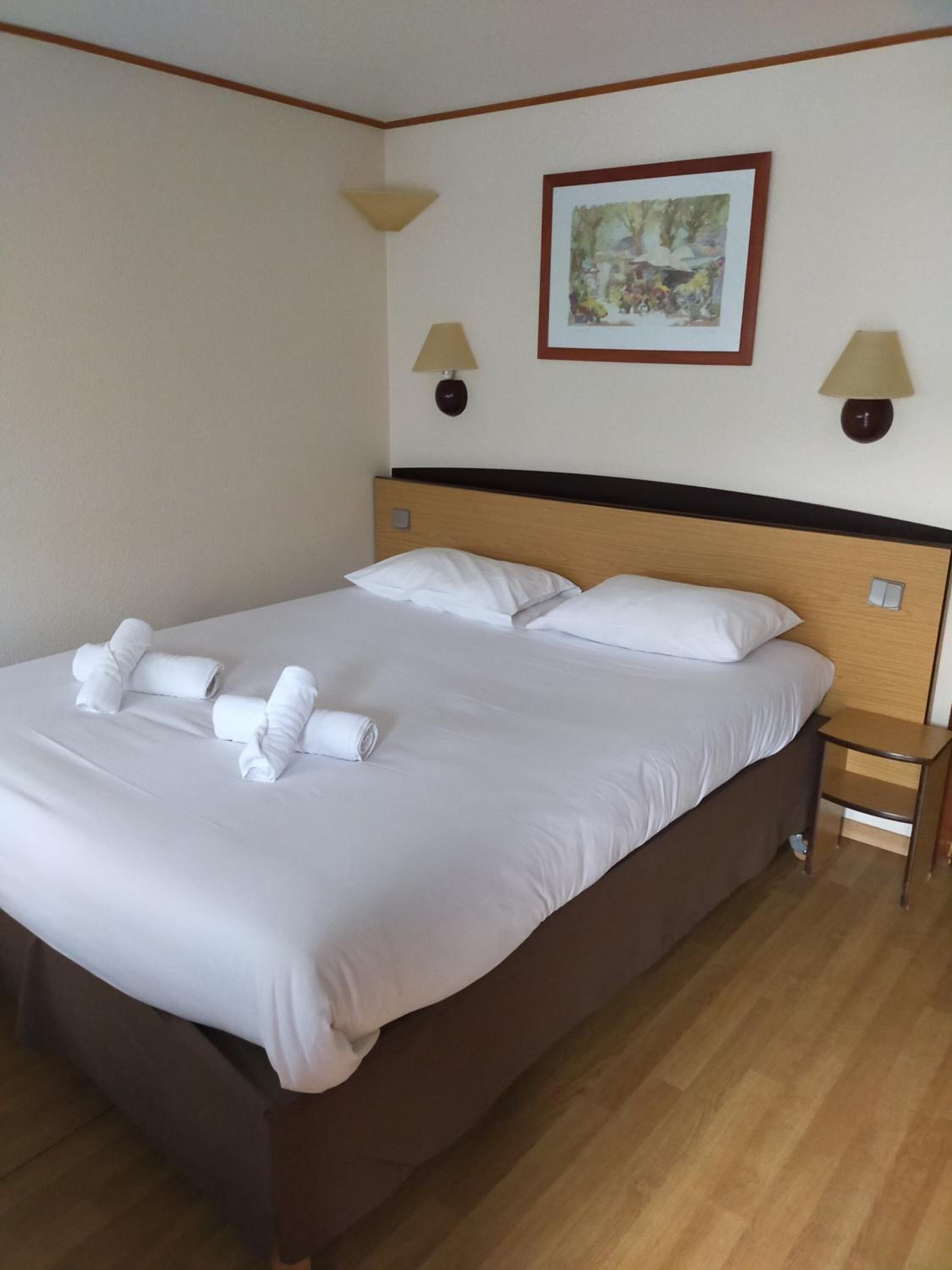 Un lit bien fait, avec du linge de lit blanc et des serviettes pliées, une tête de lit en bois, deux lampes de chevet et un tableau encadré au mur.