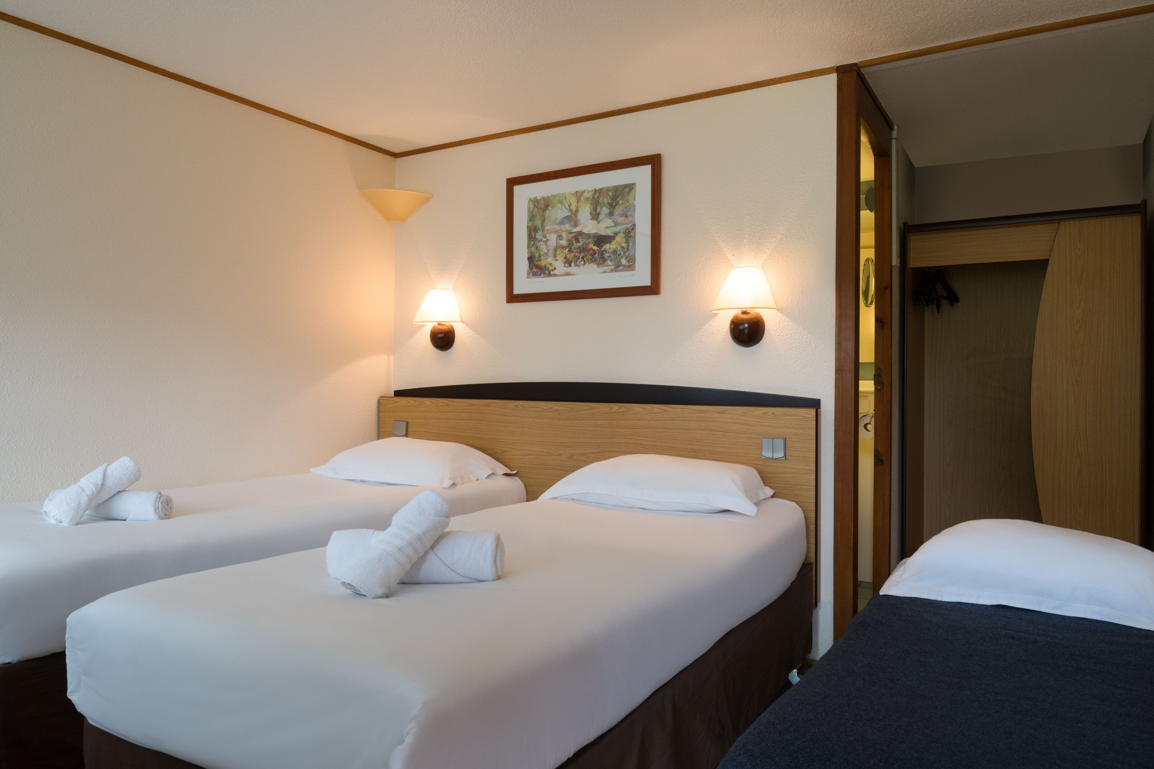 A hotel room with two beds, each with white linens and towels, a wooden headboard, and wall-mounted lamps.