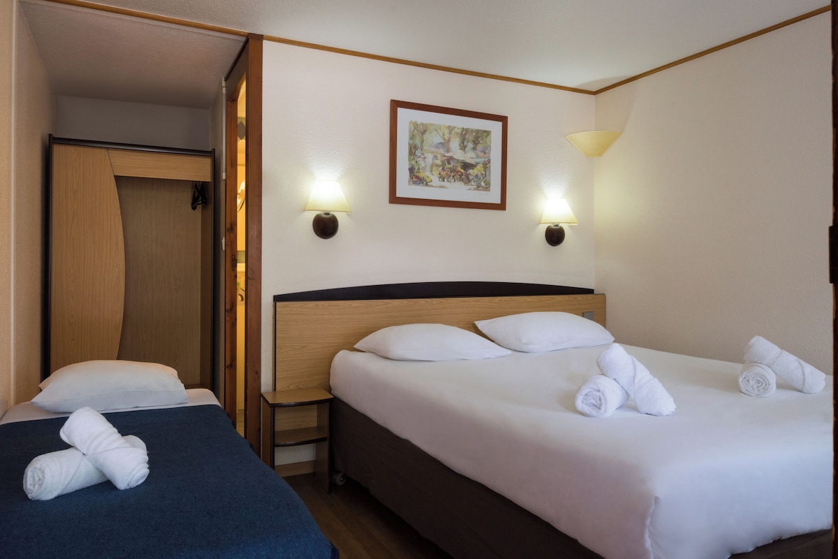 A hotel room with two beds, each with white linens and towels. There are wall-mounted lamps and a framed picture above the beds.
