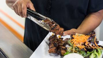 Una persona sirve brochetas de carne a la parrilla en un plato blanco, acompañadas de una ensalada y un pan blanco.