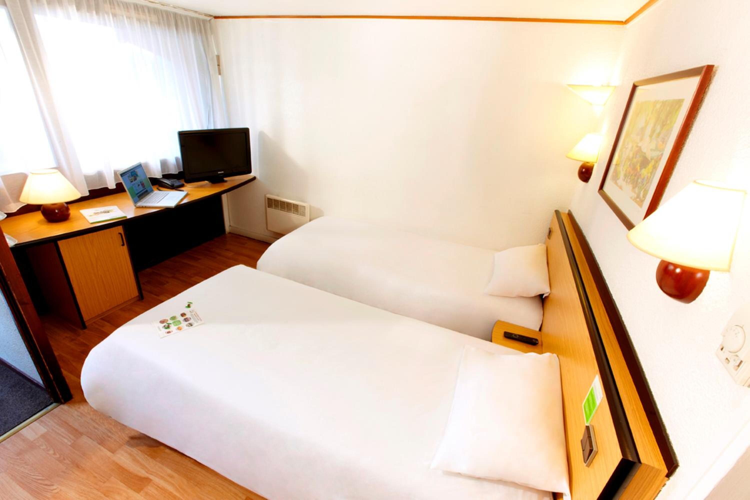 A hotel room with a bed, a desk with a laptop, a television, and a wall-mounted lamp.