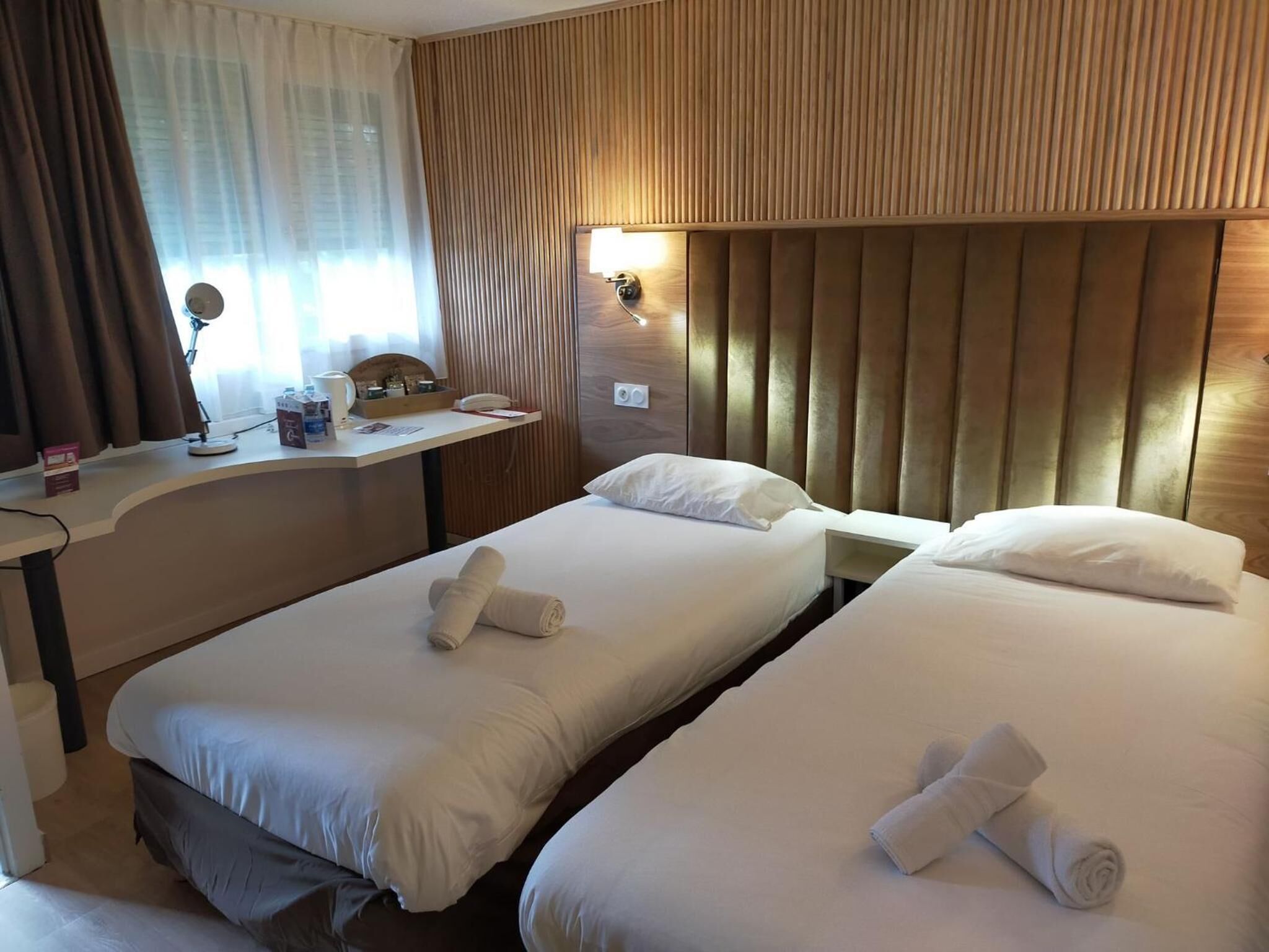 Habitación de hotel con dos camas individuales, un cabecero de madera, un escritorio con teléfono y una ventana con cortinas.