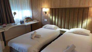 Une chambre d’hôtel avec deux lits simples, une tête de lit en bois, un bureau avec un téléphone et une fenêtre avec des rideaux.