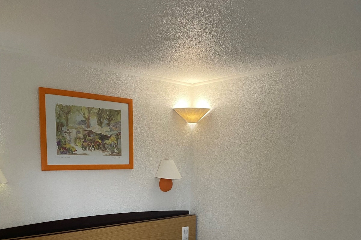 A neatly made bed with white linens, a headboard, a wall-mounted lamp, and a framed picture on the wall.