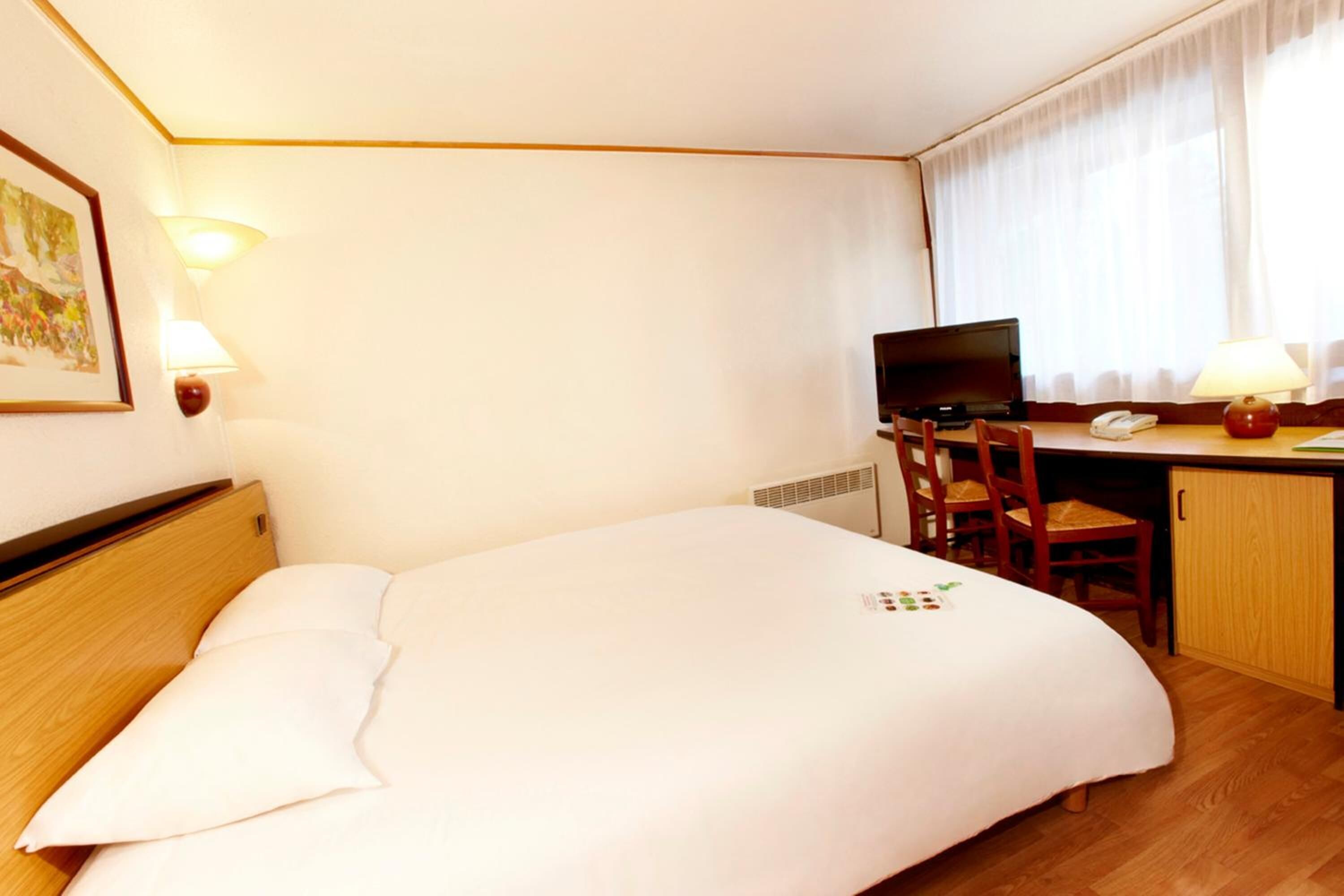 A hotel room with a bed, a desk with a computer, a chair, and a lamp.