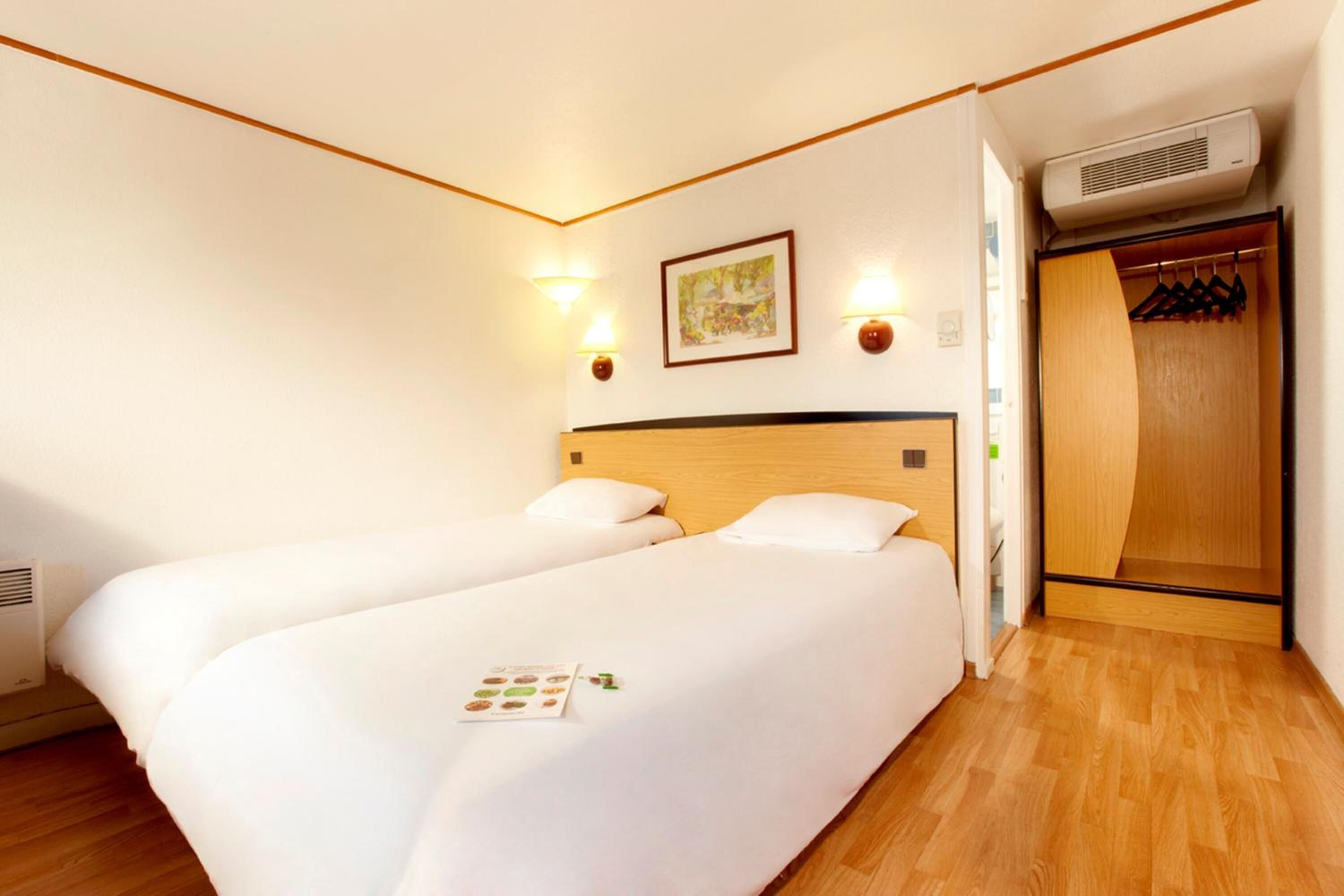 Ett hotellrum med två sängar, en sänggavel i trä, en väggmonterad luftkonditioneringsanläggning och en garderob i trä med upphängningsstång för kläder.