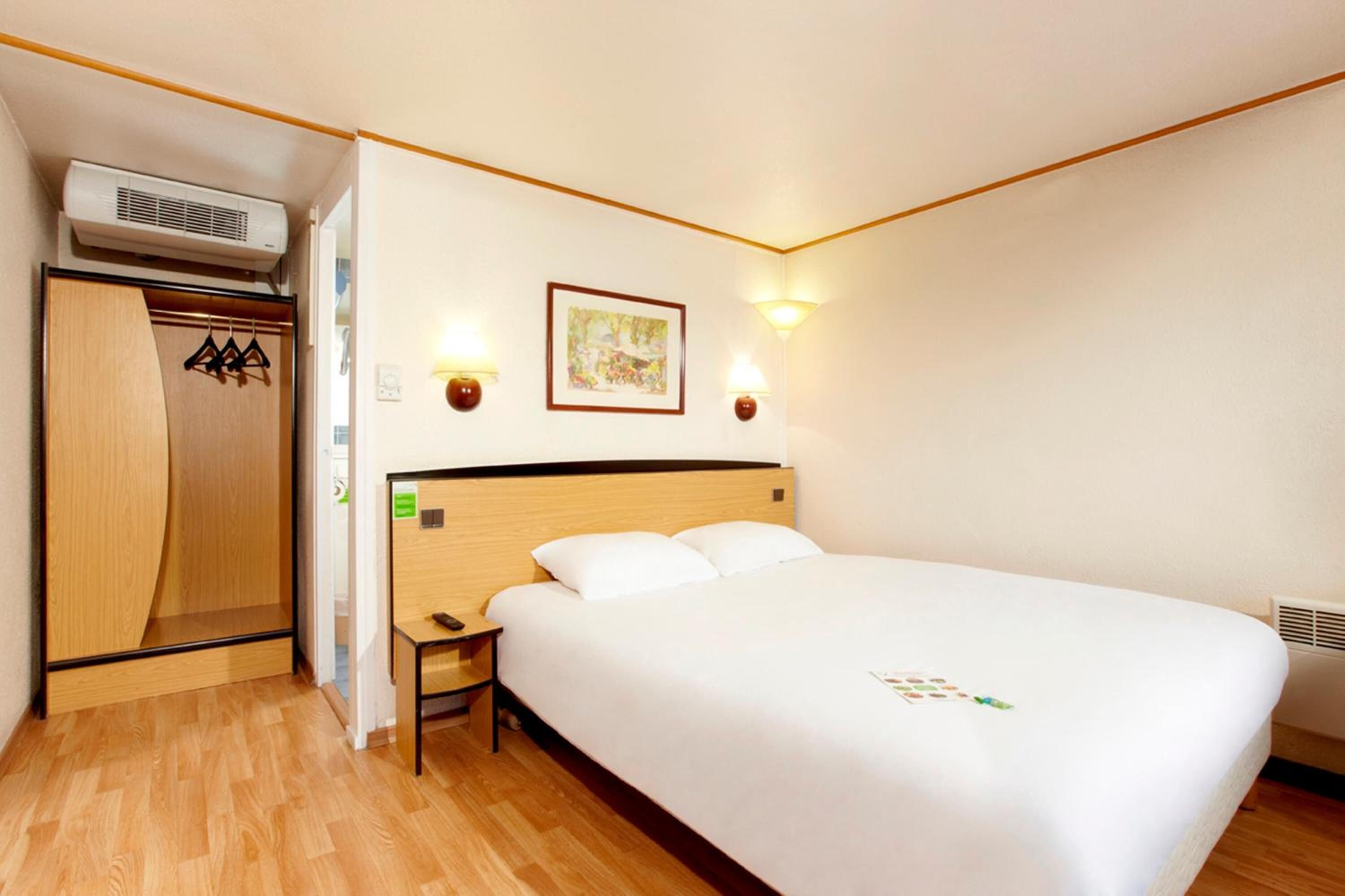 Habitación de hotel con una cama de madera, una mesita de noche, un armario y dos luces fijadas a la pared.