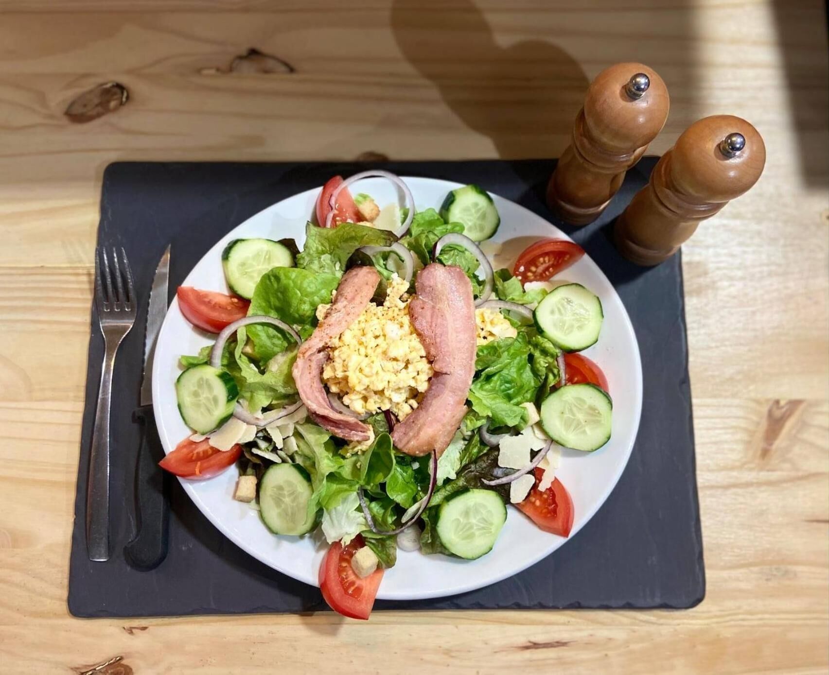 A plate of salad with cucumbers, tomatoes, lettuce, and a slice of meat, accompanied by a fork and pepper shakers on a wooden table.