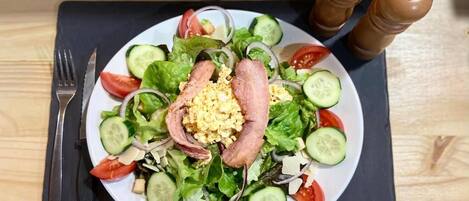 A plate of salad with cucumbers, tomatoes, lettuce, and a slice of meat, accompanied by a fork and pepper shakers on a wooden table.