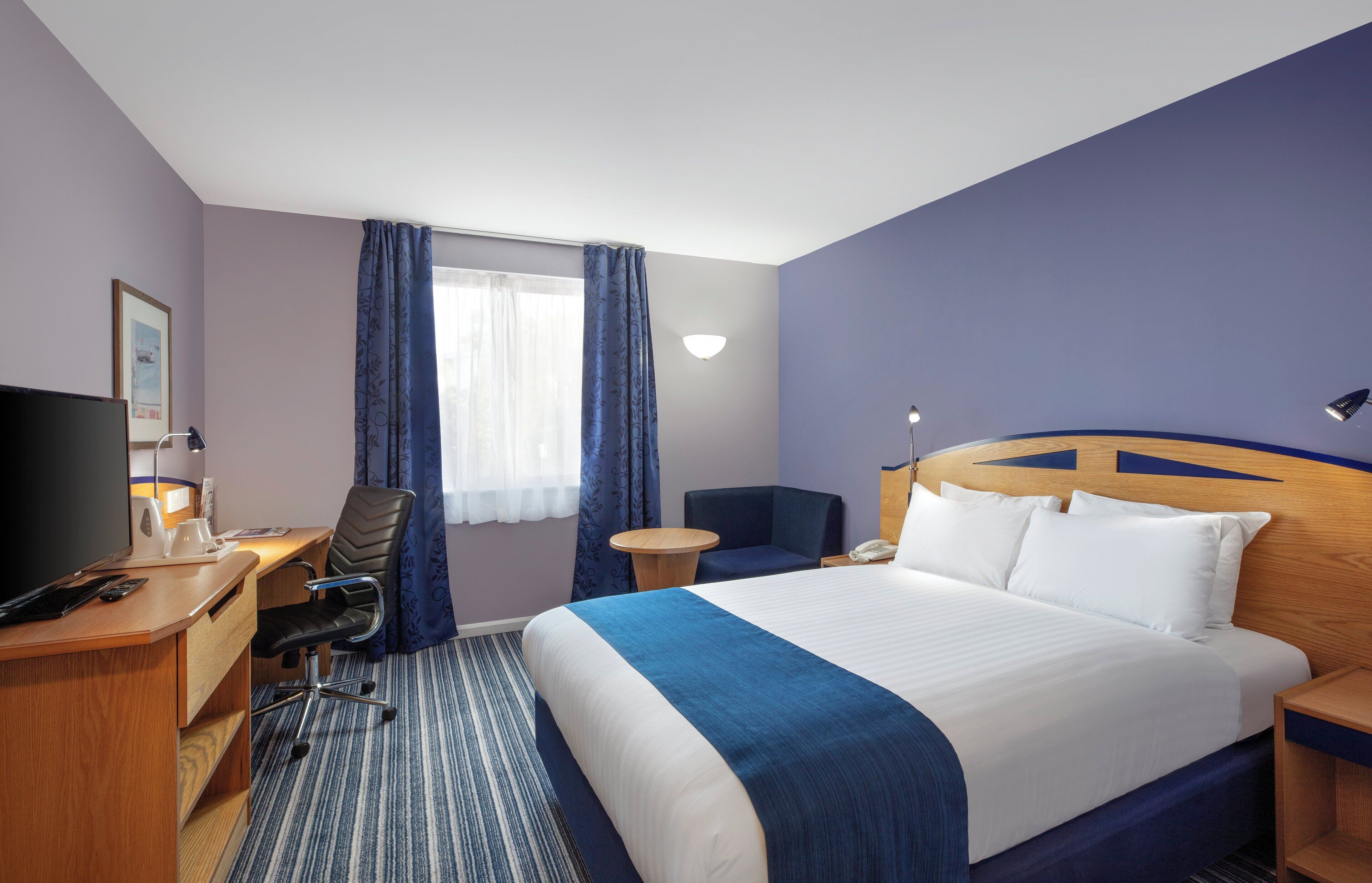 Habitación de hotel con cama, escritorio, silla, televisor y ventana con cortinas.