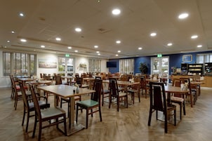 A spacious dining area with multiple tables and chairs, a kitchen area with appliances, and large windows allowing natural light.